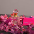 A square textured glass bottle with a pink gem top for Cherry Blossom Attar by Perfumous, styled with pink flowers.