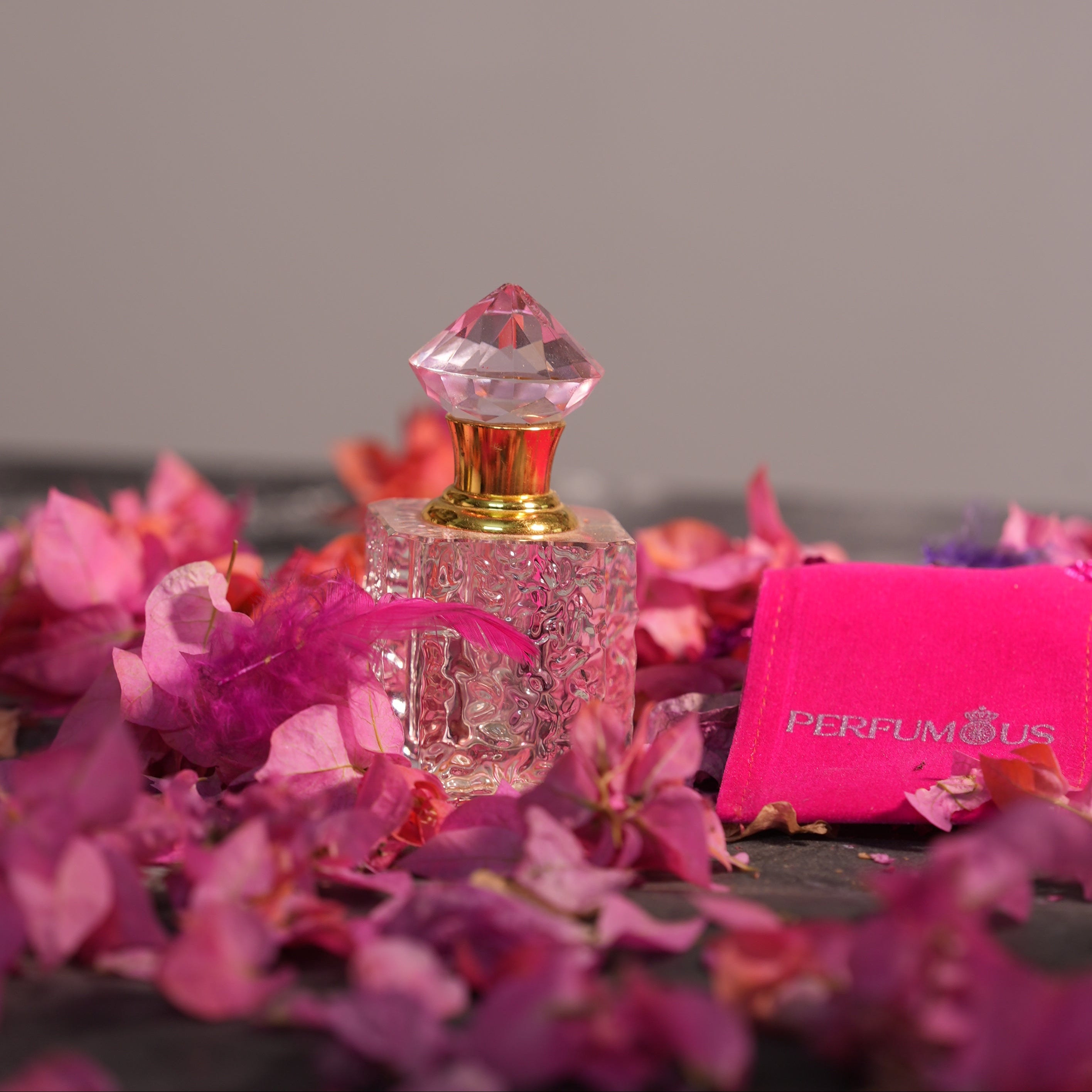 A square textured glass bottle with a pink gem top for Cherry Blossom Attar by Perfumous, styled with pink flowers.